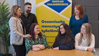 IKU-tiimin jäsenet ovat kerääntyneinä yhteen osa istuen pöydän ääressä ja osa seisten heidän takanaan hymyillen toisilleen.