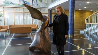 Hieman oikealla kuvassa nainen tummissa vaatteissa katselee veistosta vasemmalla. Taustalla lasia ja tiiliseinää ja ylhäällä maalattu taideteos.