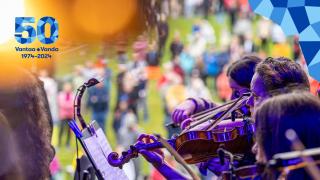 Violinists in the foreground, listeners in the park in the background. Top right corner with the corner element of the jubilee year and left corner with the jubilee year emblem.