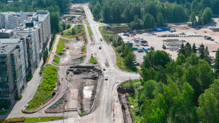 Kuvassa näkyy tie, jossa kulkee ajoneuvo-ja bussiliikennettä, työmaa-alueet ja läheinen asuinalue rakennuksineen.