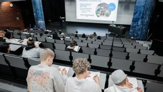 Opiskelijat istuvat auditoriossa tekemässä muistiinpanoja. Valkokankaalla näkyy Hybridikeskuksen esittelyteksti.