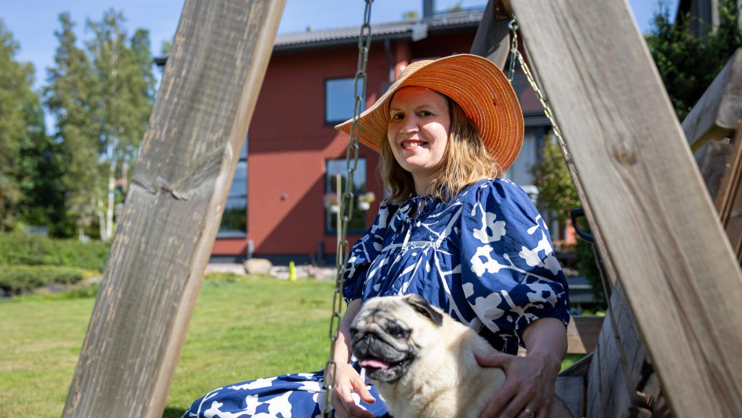 Asukas istuu hellehatussa hymyilleen pihakeinussa koiransa kanssa. Taustalla on kotitalo.