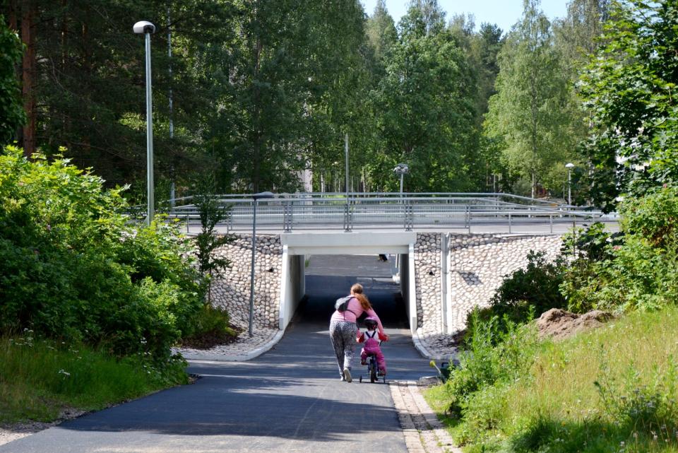 Äiti ja lapsi kävelemässä kohti siltaa kesäisessä ympäristössä. Lapsi pyöräilee ja äiti tukee lasta. 
